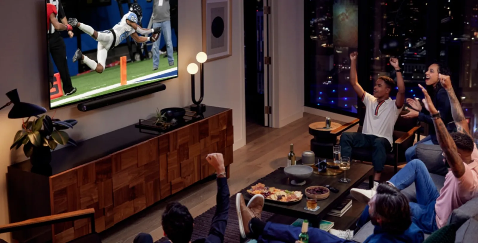 Friends cheering at a football game on a wall-mounted TV in a living room at night